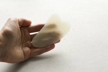 Genç bir kadın el guacha kazıyıcısını tutuyor, yüz masajı için, vücut, doğal beyaz yeşim taşı, beyaz arka plan. Güzellik ve gençlik için terapi masajı. Metnin için boşluk kopyala.