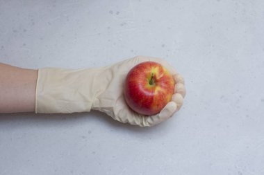 Kırmızı elma, açık gri ve beyaz modern beton arka planda beyaz, tek kullanımlık lastik eldiven içinde duruyor. Lastik eldivendeki bir el elma meyvesi tutar. Coronavirüs salgını konsepti.