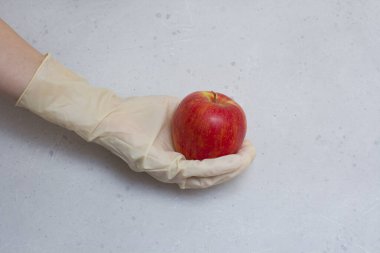 Kırmızı elma, açık gri ve beyaz modern beton arka planda beyaz, tek kullanımlık lastik eldiven içinde duruyor. Lastik eldivendeki bir el elma meyvesi tutar. Coronavirüs salgını konsepti.