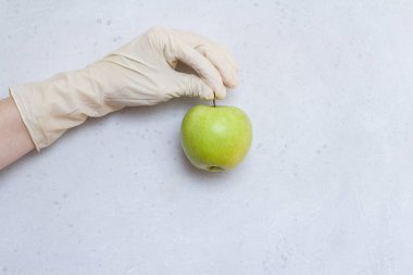 Lastik eldivendeki bir el yeşil elma meyvesi tutar. Yeşil elma, açık gri ve beyaz modern beton arka planda beyaz, tek kullanımlık lastik eldiven içinde duruyor. Coronavirüs salgını konsepti.