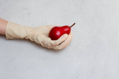 Lastik eldivendeki bir el kırmızı armut meyvesi tutar. Kırmızı armut, açık gri ve beyaz modern beton arka planda beyaz, tek kullanımlık lastik eldiven içinde duruyor. Coronavirüs salgını konsepti.