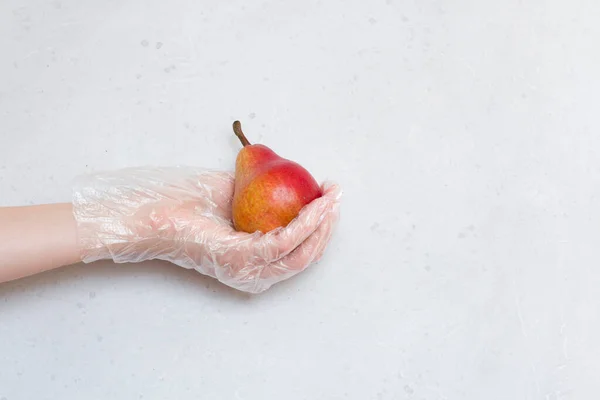 Hand in disposable transparent glove holds Red pear fruit. Red pear lies on hand in disposable transparent glove, on light gray, white modern concrete background. Coronavirus epidemic concept.