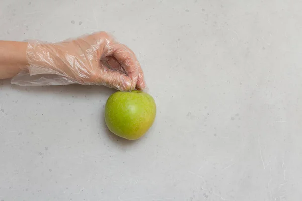 Hand in disposable transparent glove holds Green apple fruit. Green apple lies on hand in disposable transparent glove, on light gray, white modern concrete background. Coronavirus epidemic concept.
