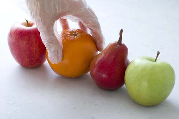 Hand in disposable transparent glove holds touches fruits of red and green apple, red pear and an orange, close-up, on a light gray and white modern concrete background. Coronavirus epidemic concept.