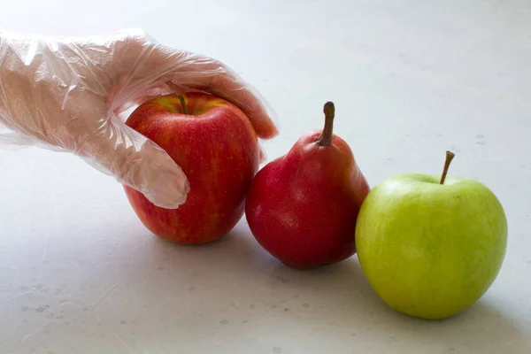 Hand in disposable transparent glove holds touches fruits of red and green apple, red pear, close-up, on a light gray and white modern concrete background. Coronavirus epidemic concept.