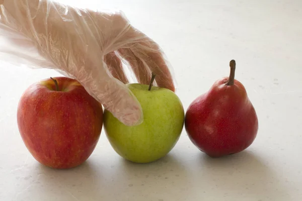 Hand in disposable transparent glove holds touches fruits of red and green apple, red pear, close-up, on a light gray and white modern concrete background. Coronavirus epidemic concept.