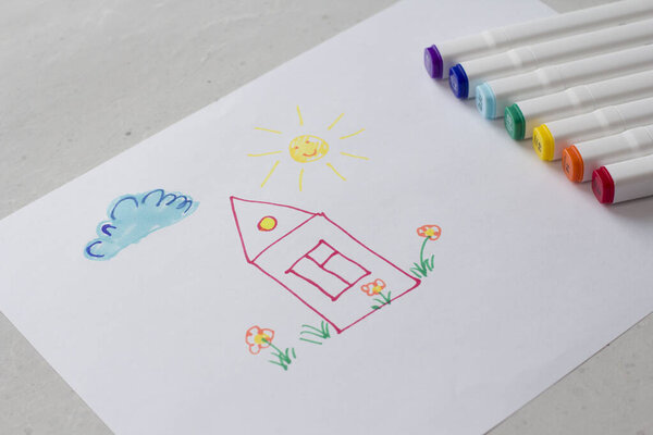 Drawn by colorful felt-tip pens a child's drawing on a white sheet of paper - a house, flowers, the sun and cloud. Creativity, drawing for children, design. Many Markers of all colors of the rainbow.