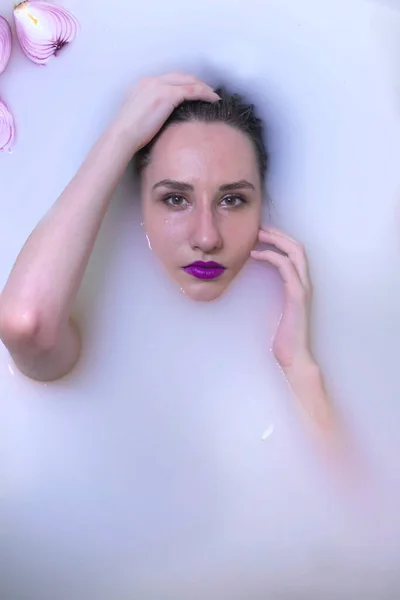 Модель моды девушка с фиолетовым макияжем в молочной ванне с фиолетовыми цветами — стоковое фото