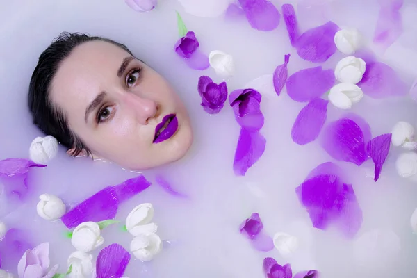 Модель моды девушка с фиолетовым макияжем в молочной ванне с фиолетовыми цветами — стоковое фото