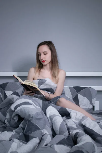 Half naakt slank blond meisje leest een mooi boek in bloemen in bed met grijze lakens in driehoeken — Stockfoto