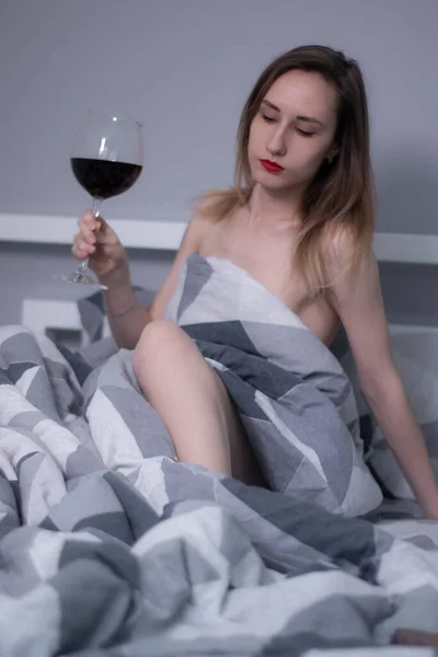 Belle fille mince à moitié nue tient un énorme verre de vin rouge au lit avec des draps gris en triangles — Photo