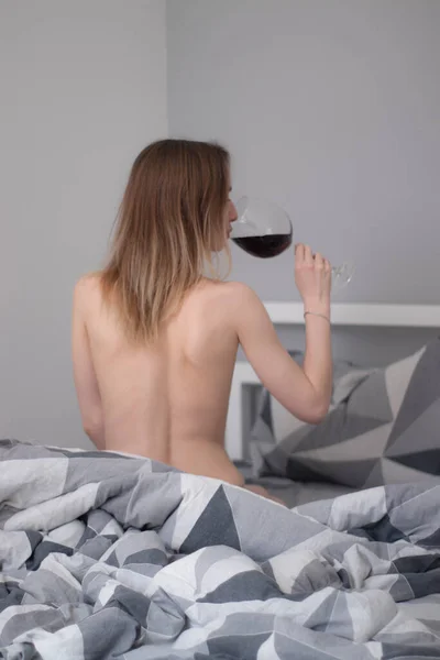 Menina seminua sentada com as costas para nós, segura um copo enorme de vinho tinto um em uma cama com lençóis cinza em triângulos — Fotografia de Stock