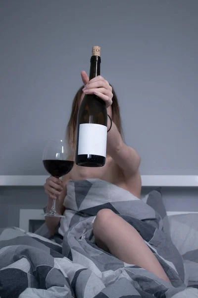 Heureuse belle fille mince moitié nue nous montre une bouteille de vin sans étiquette, au lit avec des draps gris en triangles — Photo