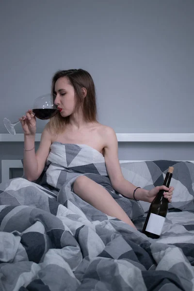 Bella snella ragazza mezza nuda beve vino rosso da un bicchiere enorme e una bottiglia senza etichetta a in un letto con lenzuola grigie in triangoli — Foto Stock