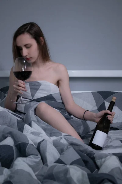 Bella snella ragazza mezza nuda beve vino rosso da un bicchiere enorme e una bottiglia senza etichetta a in un letto con lenzuola grigie — Foto Stock