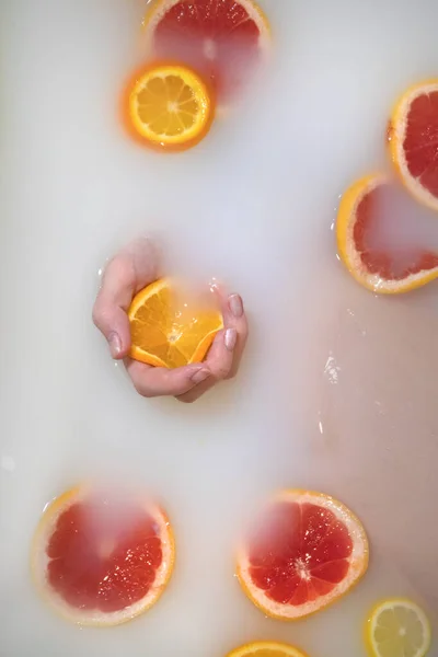 Female hand squeezes an orange in white water - milk bath. Nearby are oranges, lemons, grapefruits - vertical photo