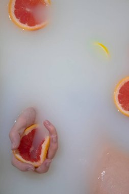 Kadın eli, beyaz suya greyfurt sıkıyor. Süt banyosu. Yakında portakallar, limonlar, greyfurtlar var.