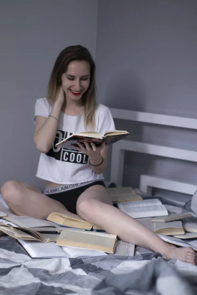 Chica delgada sentada en la cama con un montón de libros, lee. Enseñanza en casa. Estudiando en cuarentena. Leer es útil —  Fotos de Stock