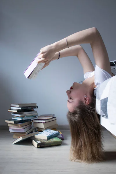 Κορίτσι που διαβάζει ένα βιβλίο ξαπλωμένο στο κρεβάτι ανάποδα με ένα σωρό βιβλία εδώ κοντά. Μαθήματα κατ 'οίκον. Μελέτη σε καραντίνα. — Φωτογραφία Αρχείου
