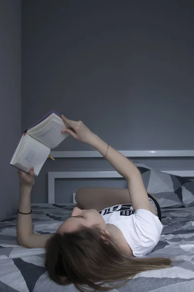 Ragazza che legge un libro sdraiata sul letto con un mucchio di libri nelle vicinanze. Studia a casa. Studiare in quarantena . — Foto Stock