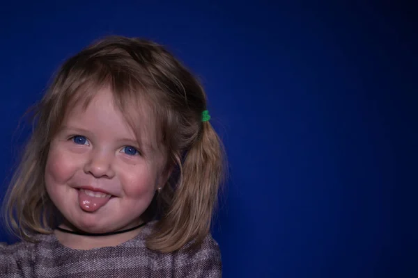 Retrato de feliz uma menina loira de três anos de idade com olhos azuis mostrando sua língua em um fundo azul — Fotografia de Stock