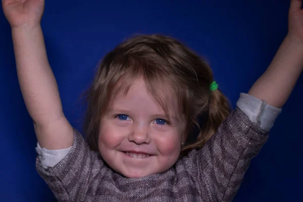 Portret van gelukkig een drie jaar oud blond meisje met blauwe ogen op een blauwe achtergrond - handen omhoog — Stockfoto