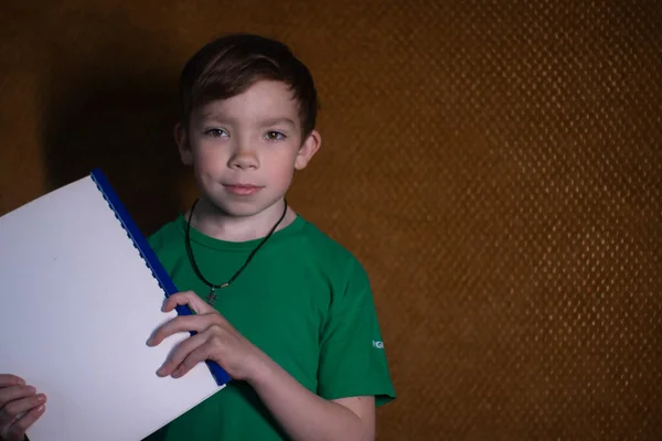 Retrato de um menino loiro de nove anos de idade em uma camiseta verde no fundo marrom segurando um caderno branco. Ensino em casa . — Fotografia de Stock