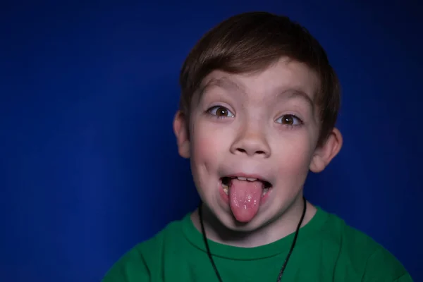 Portrét devítiletého blonďáka, který vyplázl jazyk na modrém pozadí — Stock fotografie