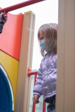 Tıbbi Tek Kullanılabilir Yüz Maskesi takan sevimli sarışın bir kızın portresi. Karantina sırasında oyun parkında oynuyor. Evde kal. İzolasyon. Coronavirus ve Hava kirliliği kavramı.