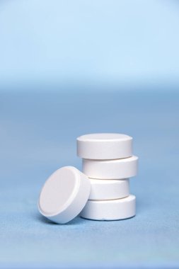 Mavi desenli yüzeyde dikey yığınlar halinde gruplandırılmış büyük beyaz C vitamini tabletlerinin yakın çekimi. Bir tablet yığının en altında. Seçici odaklanma. Kopyalama alanı olan dikey arkaplan.
