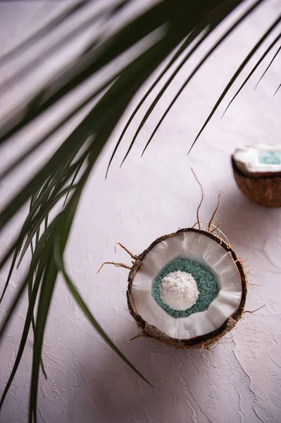 Two halves of broken coconut in its hairy brown shells and unfocused green palm leaf on white textured surface. Coconuts are filled with blue coconut flakes and white coconut candy. Selective focus.