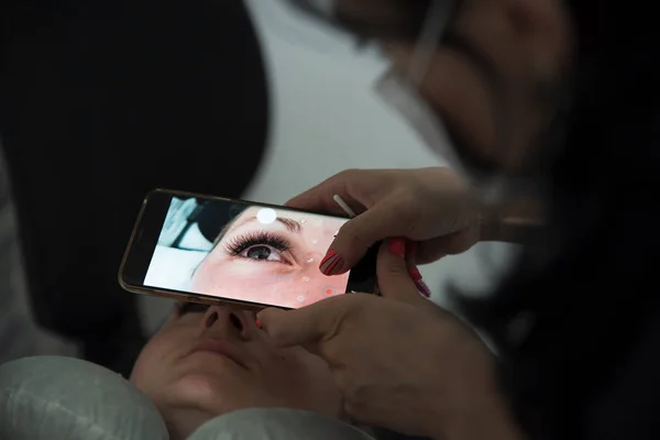 Kosmetolog robi zdjęcie za pomocą smartfona i poprawia wynik zabiegu przedłużania rzęs w salonie kosmetycznym. Samopromocja i reklama prywatnych przedsiębiorstw w Internecie — Zdjęcie stockowe
