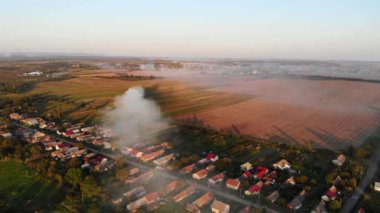 Tarlaların ve köyün üzerinde sigara iç. Gün batımında kırsal alan manzarası. Sonbaharın başlarında, hasat zamanı. Kirlilik ve ekoloji teması. Hava Aracı 4K Sinematik Görüntüler