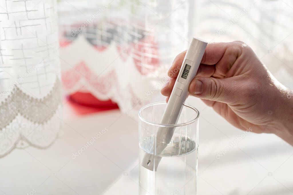 Process of checking the purity level of tap water using water TDS meter tool, close up. Device shows 198 ppm of contaminant level, which is considered the typical value. At home in the kitchen.
