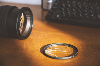 Fotoğrafçı çalışma masasındaki DSLR kamerasının yanındaki kırık UV filtresi. Ezilmiş bir filtre ve kaydedilmiş bir lens örneği. Pahalı bir lensi hasardan koruma teması. Matte etkisi.