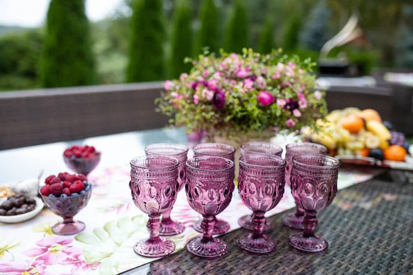 Piękne szklanki kolorowego fioletowego szkła na stole z jagodami i kwiatami — Zdjęcie stockowe