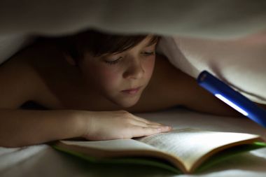 Genç çocuk battaniye veya yorgan altında bir kitap okuma