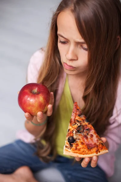 Заботливая молодая девушка обдумывает выбор фруктов или кусочка пиццы — стоковое фото