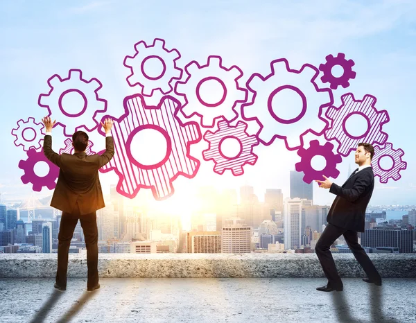 Young businessmen in suits holding abstract cogwheel sketch on rooftop with city view and sunlight. Teamwork concept — Stock Photo, Image