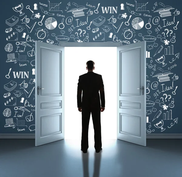 Back view of young businessman standing in room with business icons on wall against open door with bright light. Success concept — Stock fotografie