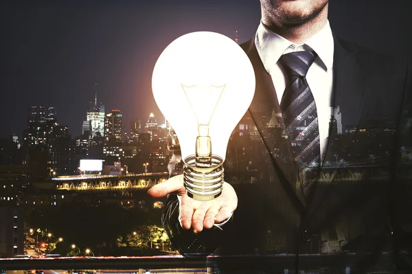 Businessperson in suit and tie holding large light bulb on city background. Idea concept. — Stockfoto
