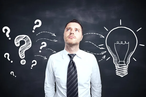 Handsome young male thinking on blackboard background with creative light bulb and question marks sketch. Idea concept — Stock Photo, Image