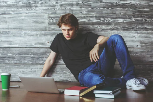 Vonzó fiatal srác ül a fa padló a könyvek, a csésze kávé és a laptop. Oktatási koncepció — Stock Fotó
