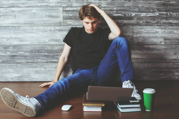 Joli jeune homme assis sur le sol en bois avec des livres, une tasse de café et un ordinateur portable. Concept d'éducation — Photo