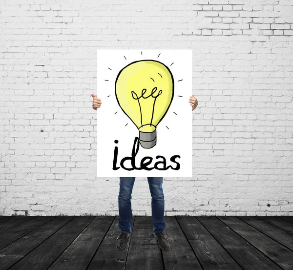 Young man in white brick room holding poster with creative lamp drawing. Idea concept — Stock Photo, Image