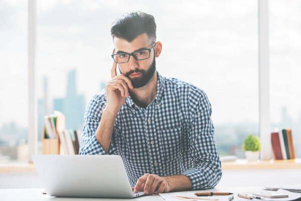 Portrait of handsome young businessman using laptop at modern workplace with blurry city view. Project concept