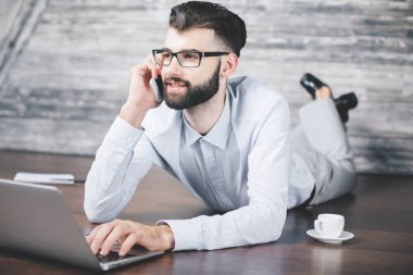Kahve fincanı ile ahşap yerde yatarken, telefonda konuşurken ve dizüstü bilgisayar kullanarak yakışıklı beyaz adam. İletişim kavramı