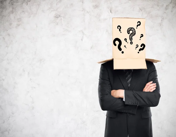 Businessman with folded arms and cardboard box with question marks covering head on concrete background. Confusion concept — Stock Photo, Image