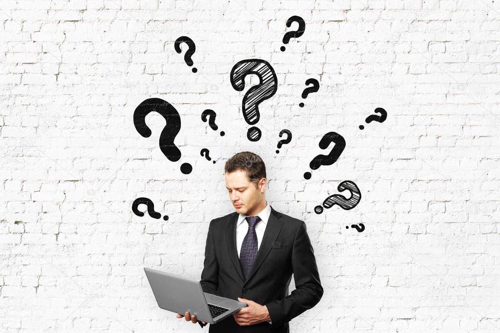 Businessman with laptop question marks on brick wall background. Confusion concept
