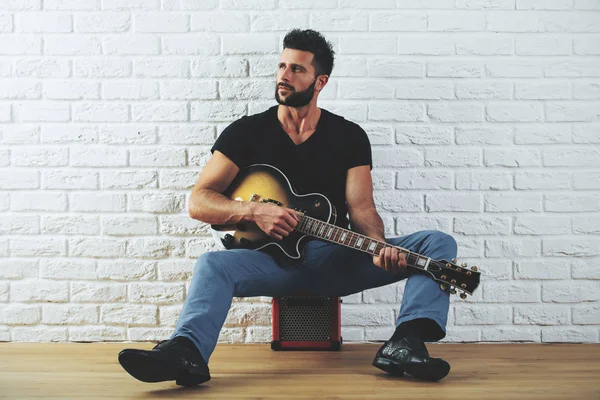 Gutaussehender europäischer Mann in lässiger Kleidung, der Gitarre spielt, während er im Innenraum mit weißer Ziegelwand und Holzboden steht. Kunstkonzept — Stockfoto